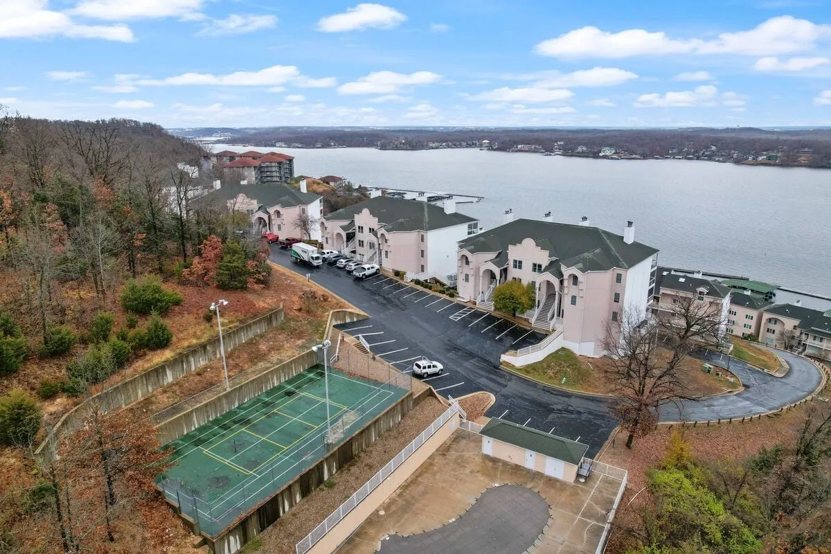 A well-maintained tennis court sits adjacent to the shimmering lake, framed by residential buildings in the background. The open, airy setting invites both recreational play and tranquil moments by the water.