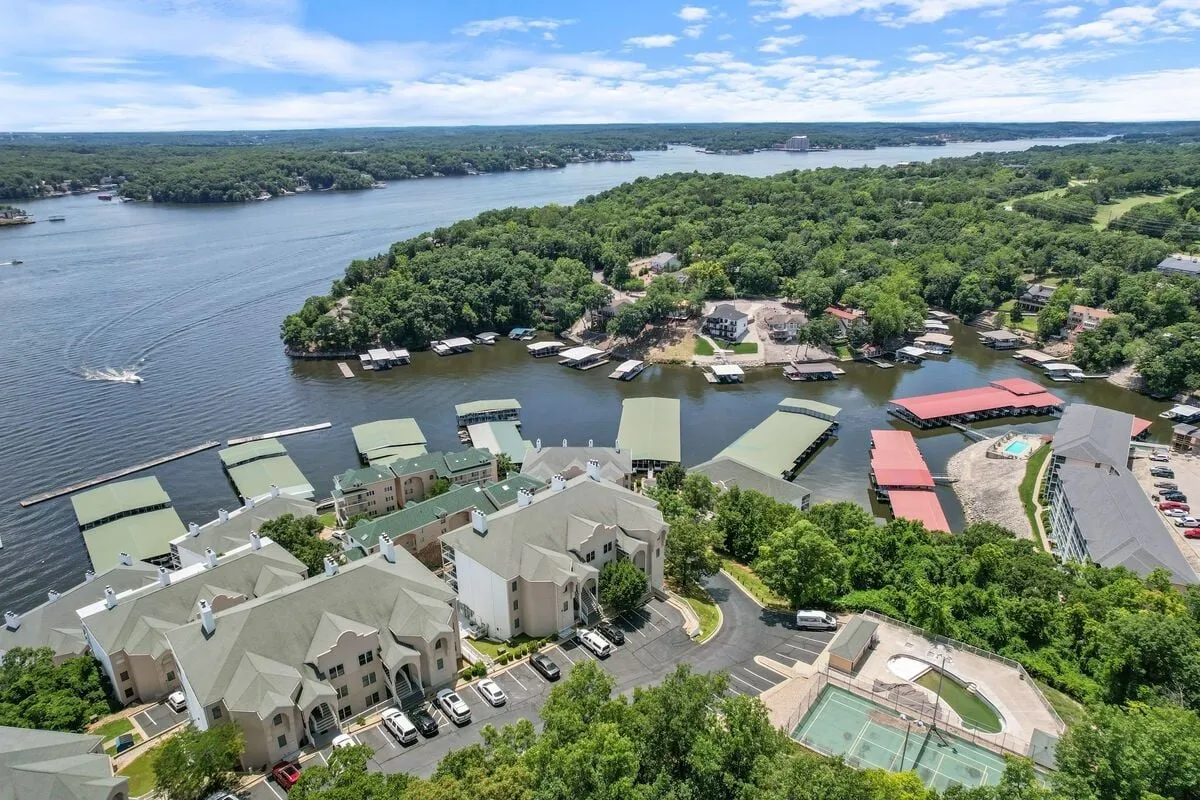 A panoramic view of the Lake Ozark waterfront, with lush greenery, marinas, and residential areas visible. The serene scene includes boats and waterfront activities, capturing the vibrant local life and appeal of the area just beyond the home.