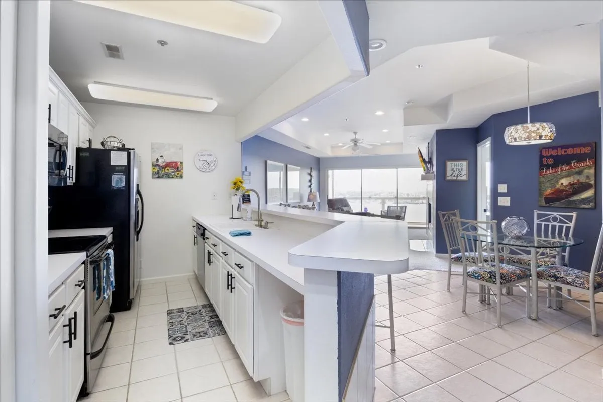A stylish kitchen and dining area featuring a kitchen island, a spacious dining table, large windows that frame the lakeside view, and decorative wall art. The space feels welcoming and ideal for sharing meals or hosting gatherings, with sunlight streaming in during sunset.