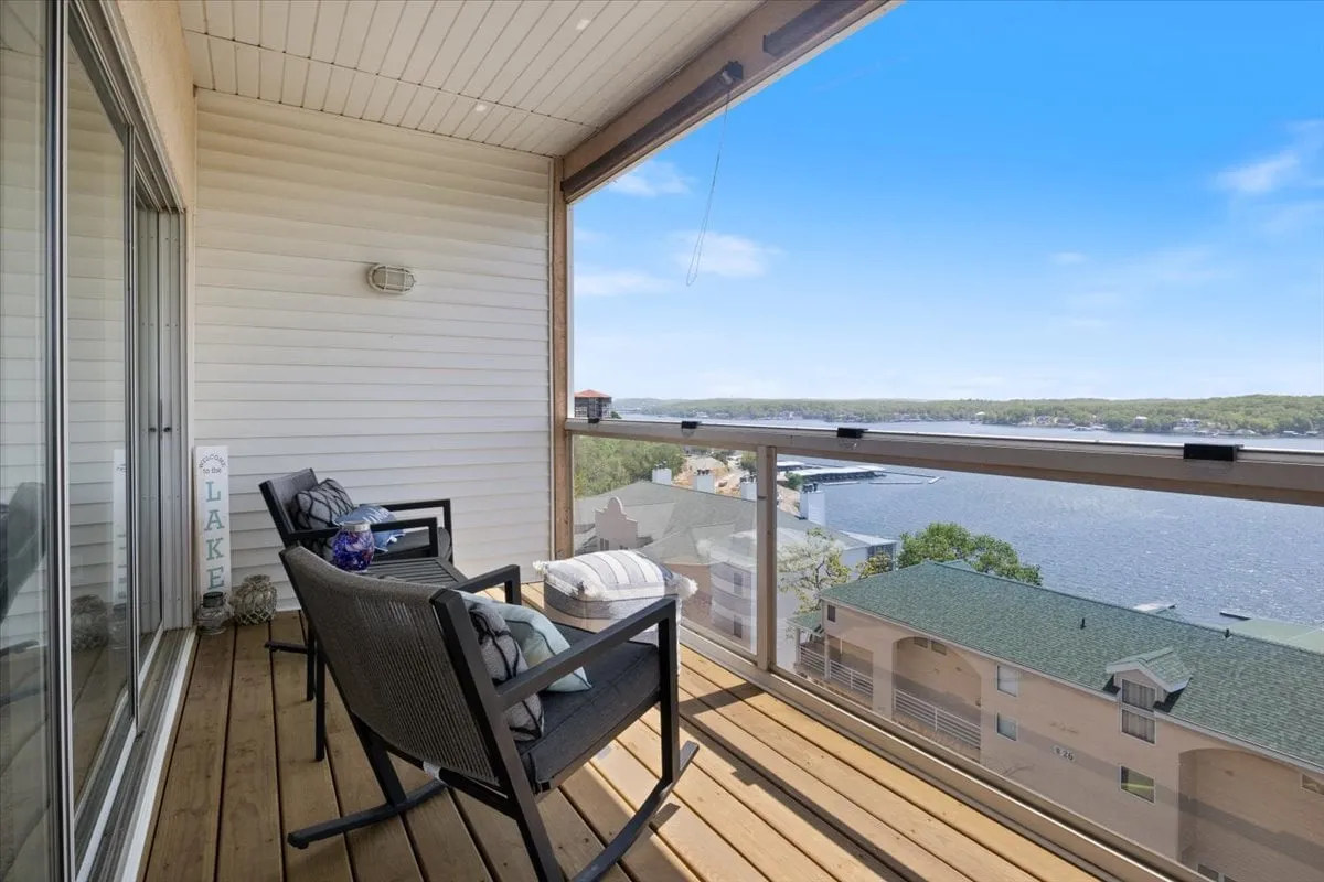 An inviting wooden deck furnished with outdoor seating overlooks the lake. The view captures water, boats passing by, and a tranquil sky, reinforcing the 'perched above the shimmering lake' feeling described in the section.
