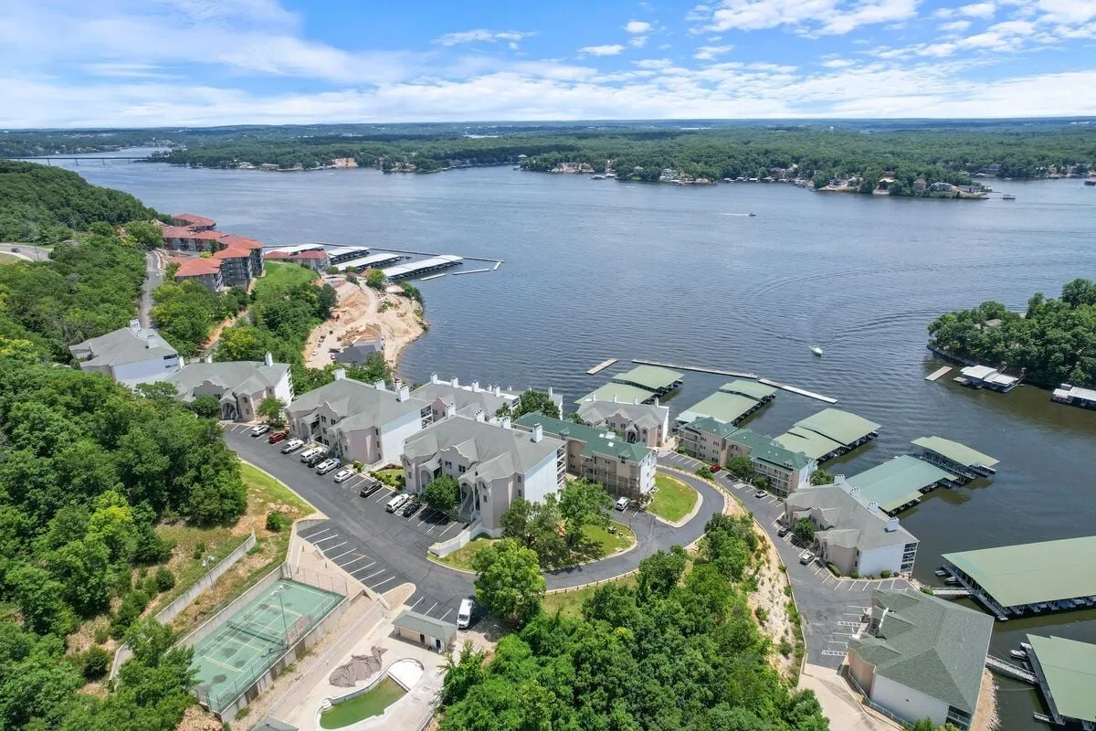 A sweeping view of the lake with boats docked at marinas, surrounded by lush greenery and residential buildings. The image captures both the waterfront activity and the peaceful environment, directly highlighting access to boating, fishing, and water sports.