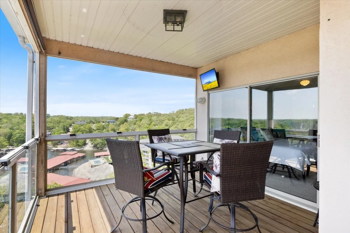 A casual outdoor dining area on a balcony, complete with a table for group meals and a scenic lake view in the background. A television mounted on the exterior wall adds an element of entertainment, making this space ideal for socializing or winding down after an evening out.