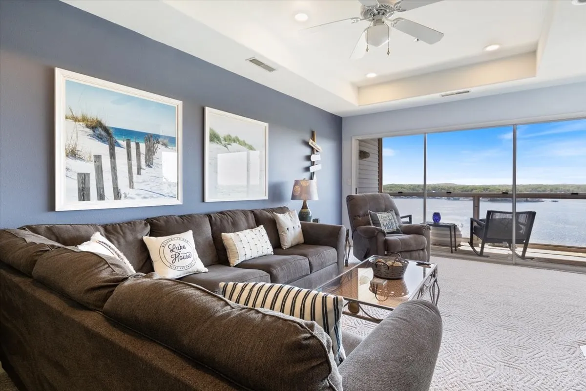A bright living room with large windows directly overlooking the water, comfortable seating including a plush sofa, and cheerful beach-themed artwork. The sunlight streams in, highlighting the tranquil, open atmosphere and the connection between indoors and the scenic lake outside.