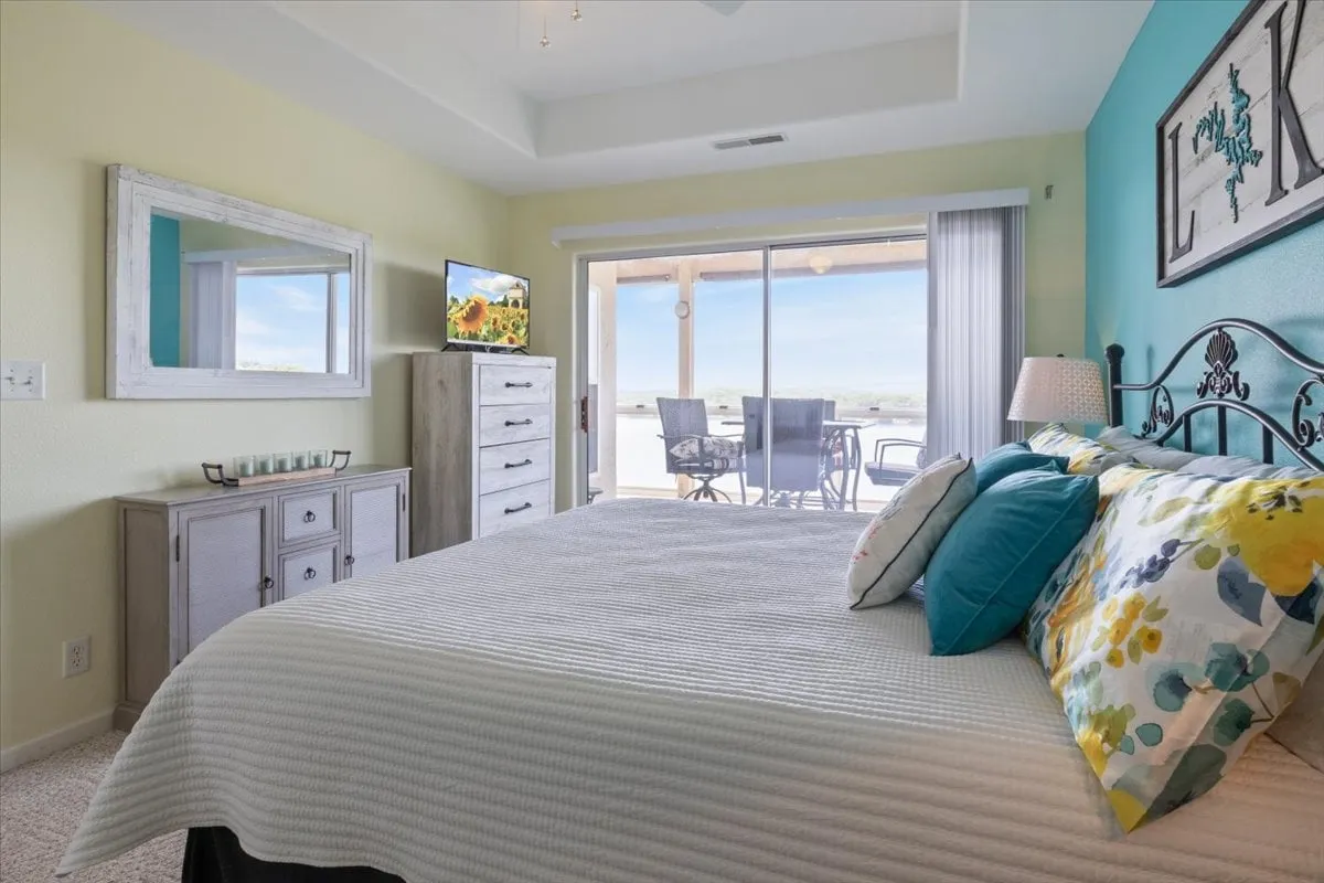 A serene master bedroom featuring a bed with colorful pillows, a large sliding glass door that opens to a balcony or patio, a dresser with a TV, and decorative wall art. The room is filled with natural light and offers views of the outdoors, suggesting a tranquil lakeside retreat.