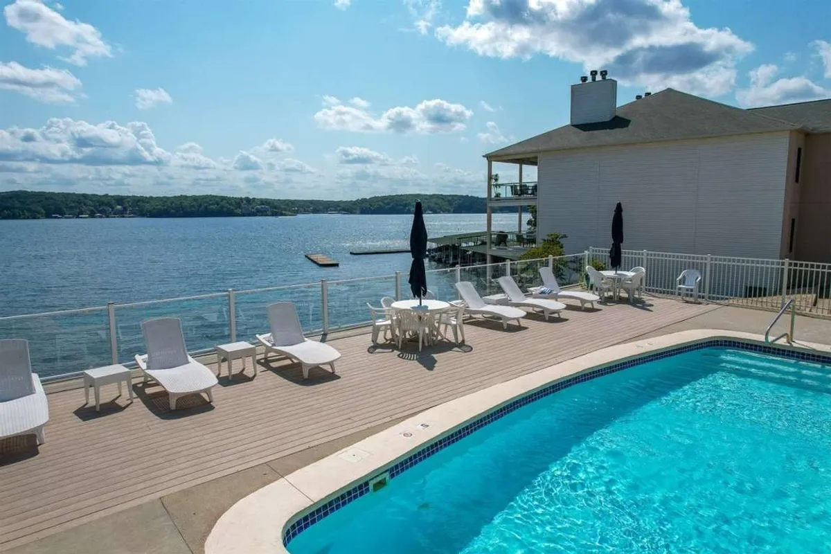 A sparkling outdoor swimming pool is set against a scenic lake view, with inviting deck chairs arranged poolside and a building structure nearby. The tranquil atmosphere hints at leisurely swims, sunbathing, and relaxation by the water.