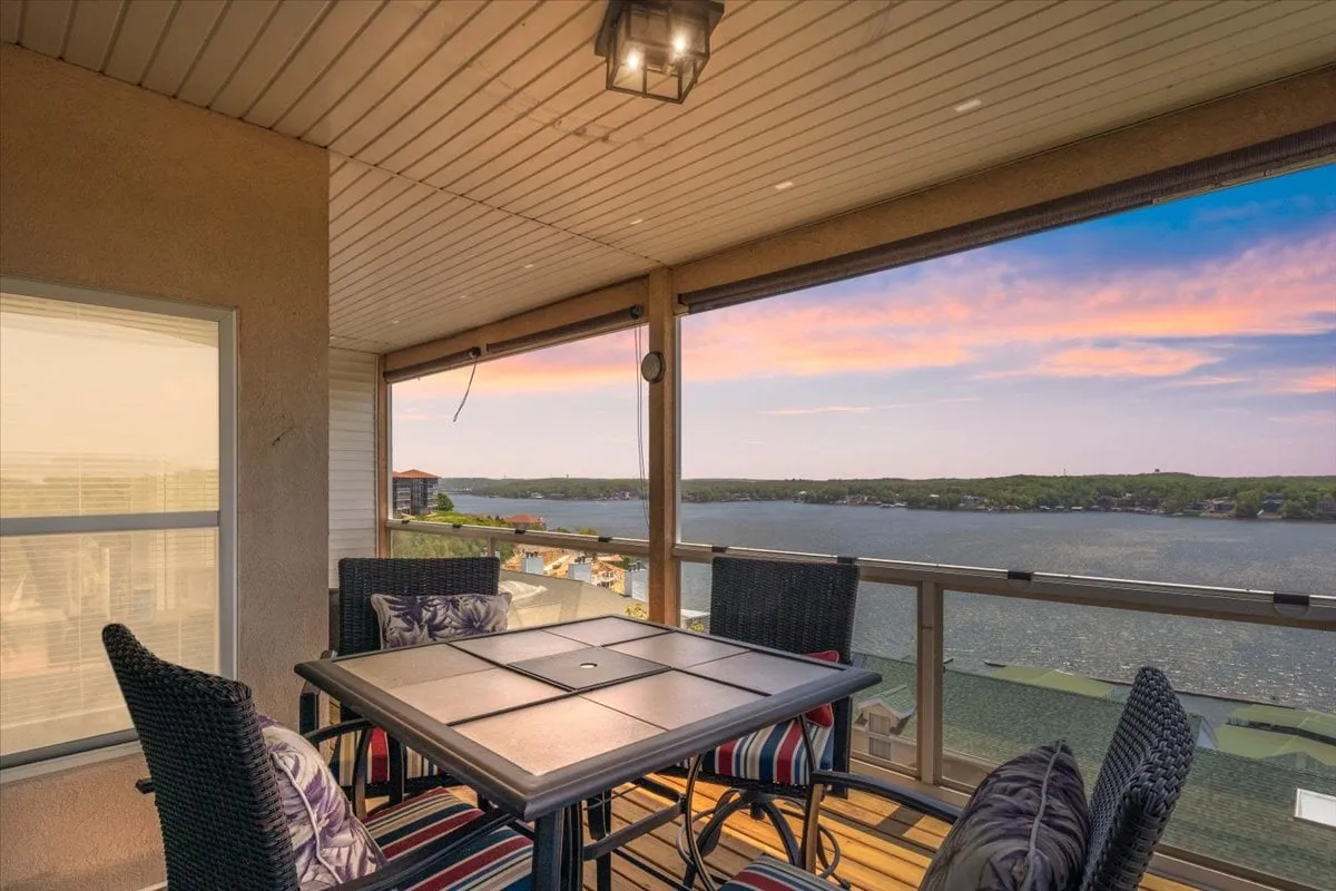The image showcases a stylish outdoor dining area on a new cedar deck, featuring striped chairs around a table, all set against a sweeping panoramic backdrop of Lake Ozark. The sky is tinged with the warm colors of sunset, inviting guests to relax, dine, and socialize while overlooking the tranquil water.