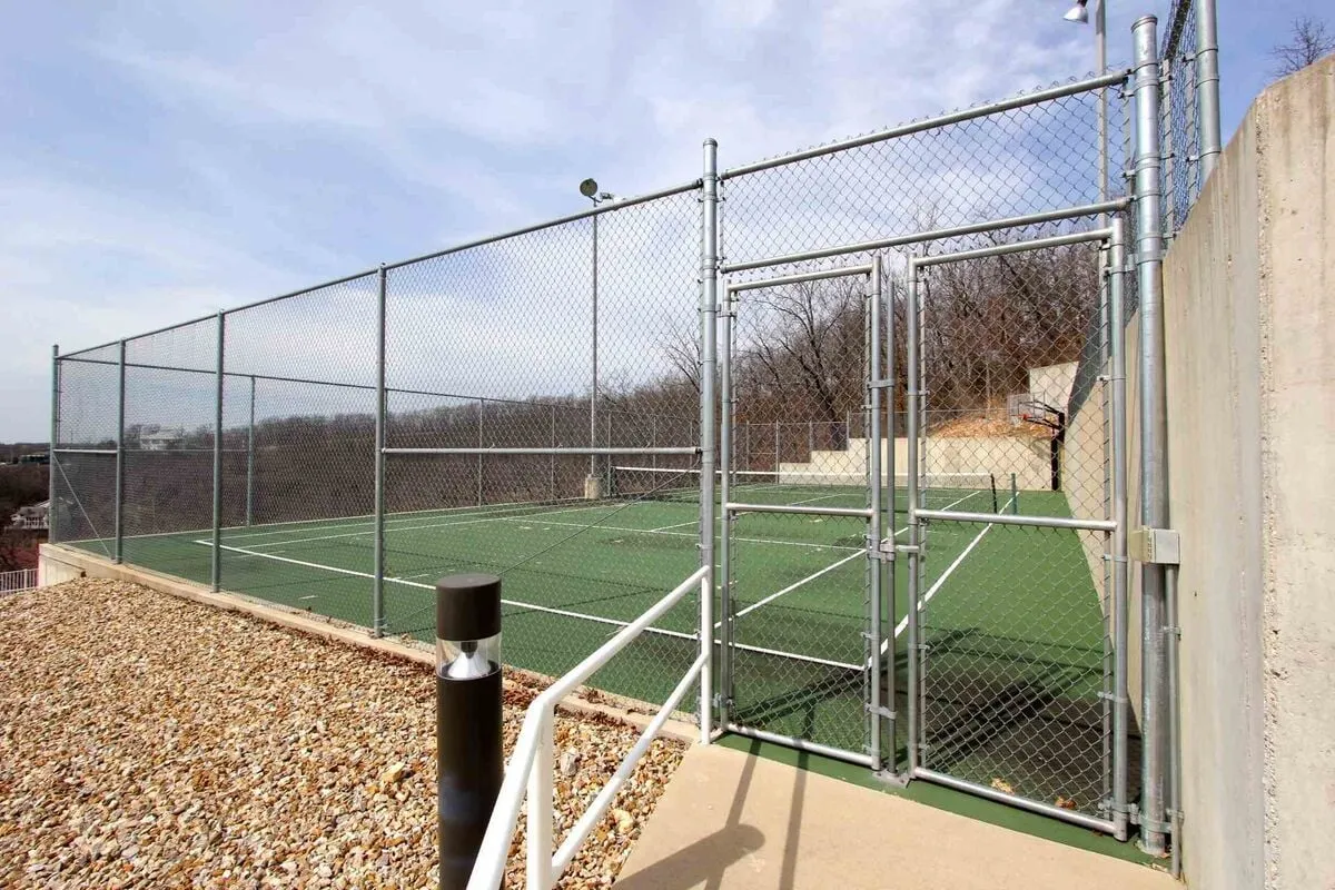A well-maintained tennis court within a fenced area, with an inviting entrance gate. The court is ready for recreational play and sports, set against a backdrop of green surroundings.
