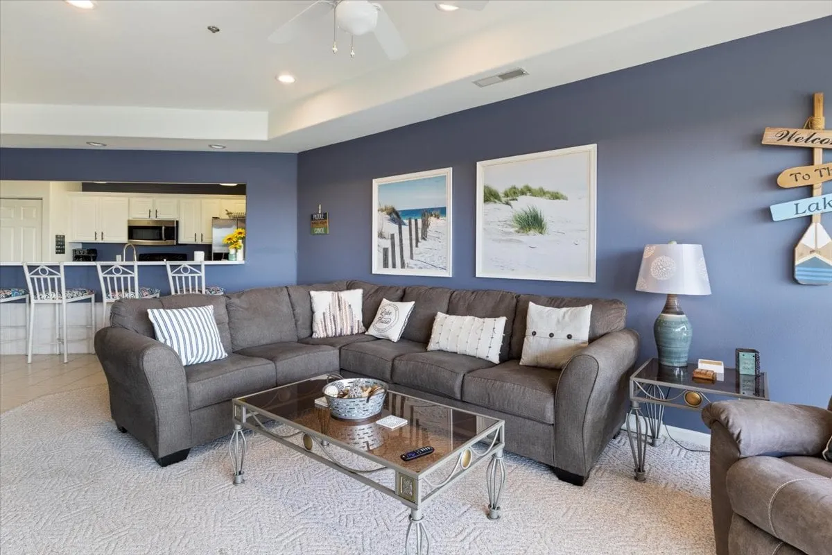 A cozy open-concept space with a large sectional sofa, glass coffee table, creative wall art, and a visible kitchen bar area in the background. The arrangement suggests an inviting spot for movie nights, board games, or gathering with friends and family.