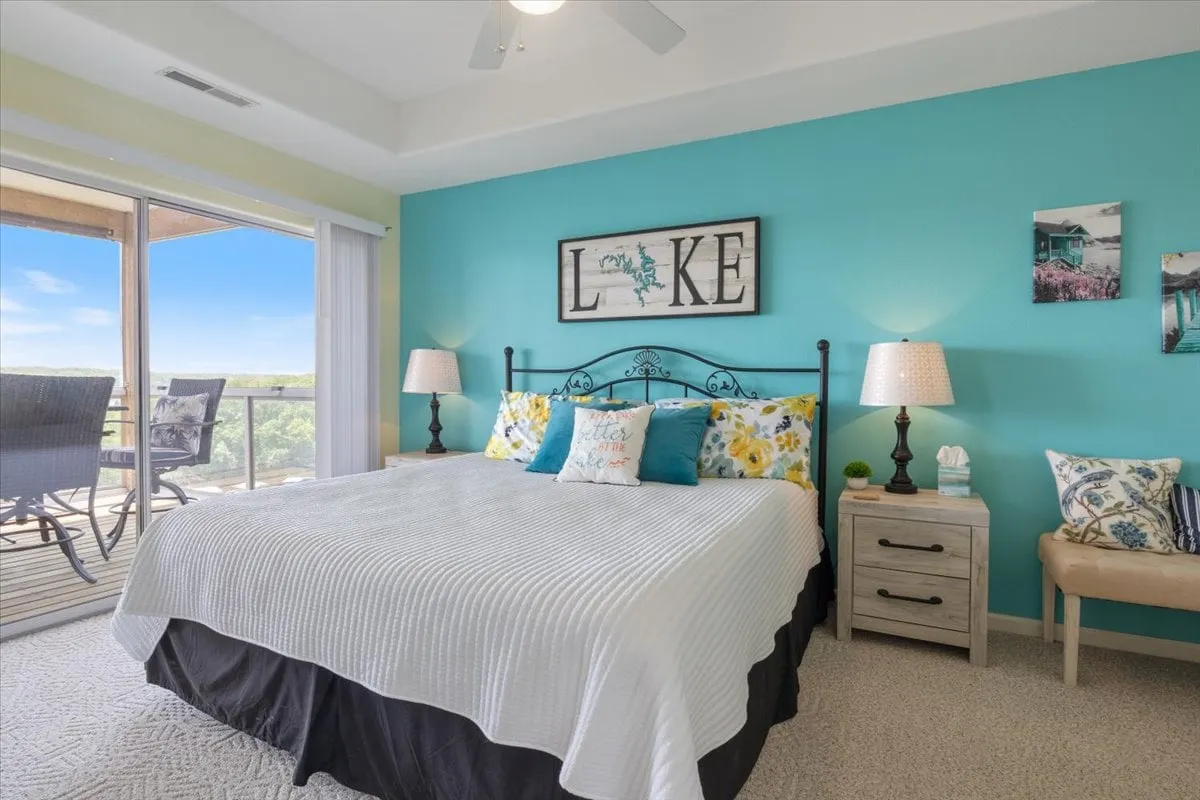 A stylish bedroom showcasing a bed with decorative pillows, wall art that says 'LAKE', nightstands with lamps, and a large window leading out to a balcony. The space is bright, tranquil, and perfectly positioned to capture morning light and lake views.